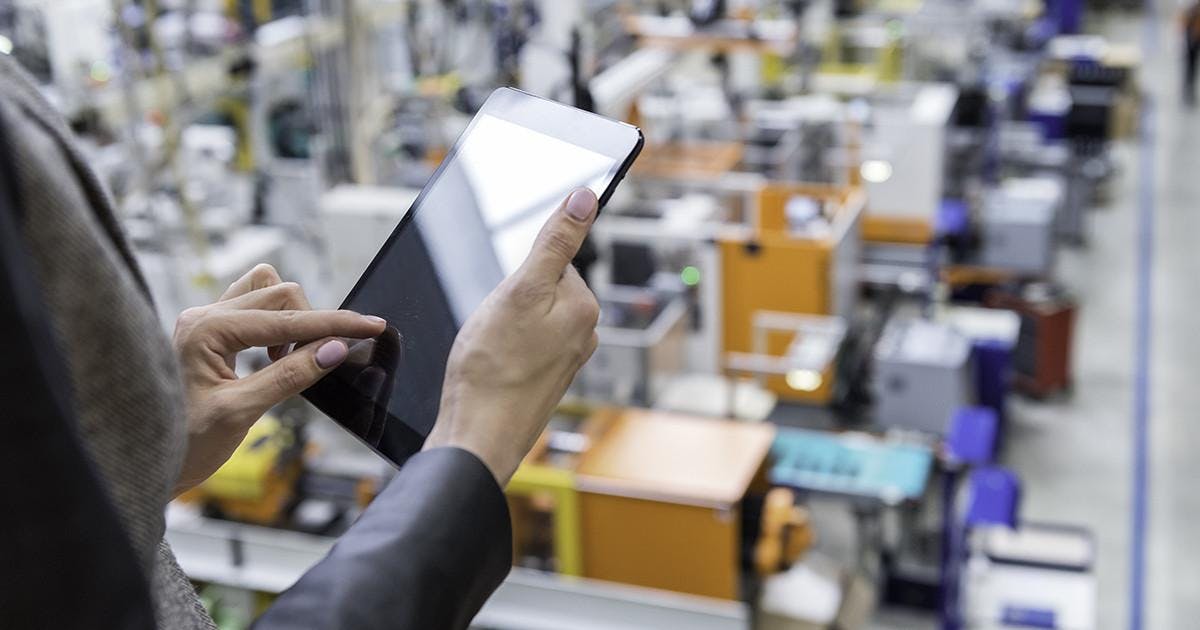 Horizontal color image of businesswoman - unrecognizable person - working with digital tablet in large futuristic factory. Woman standing on top of a balcony, holding touchpad and checking inventory of a manufacturing company on touchscreen tablet. Focus on businesswoman's hands holding black tablet, futuristic machines in background.