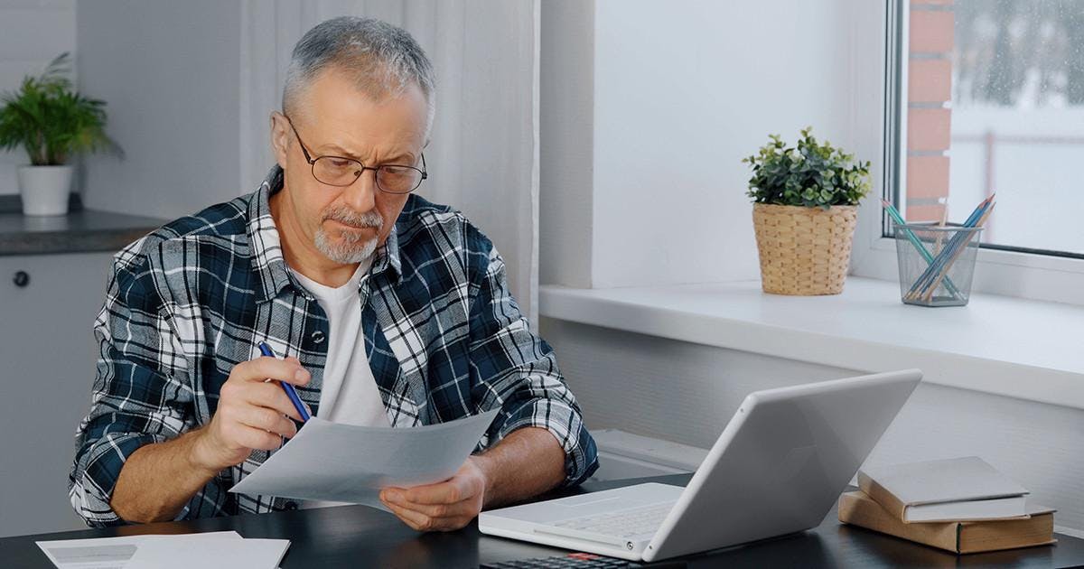 A brooding elderly man with glasses and a beard is at his laptop, looking through documents and taking notes.