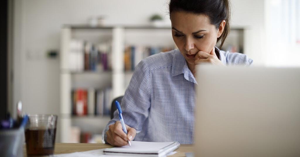 Woman working from home, writing in notebook in front of a laptop
